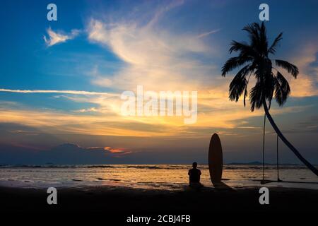 Silhouette d'homme de surf assis avec une planche de surf sur la plage. Scène de surf sur la plage au coucher du soleil avec un ciel coloré. Sports nautiques en plein air aventure style de vie. Banque D'Images