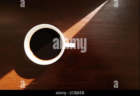 Tasse de café en céramique blanche avec café noir expresso sur fond de table en bois avec lumière du soleil triangulaire et ombre de la fenêtre, vue du dessus Banque D'Images