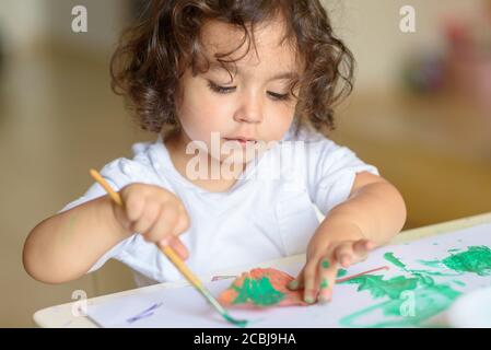 Mignon et bouclés petite fille peinture avec des peintures vertes couleur et brosse sur les vies d'automne. Dessin d'enfant adorable et maudit à la table. Banque D'Images