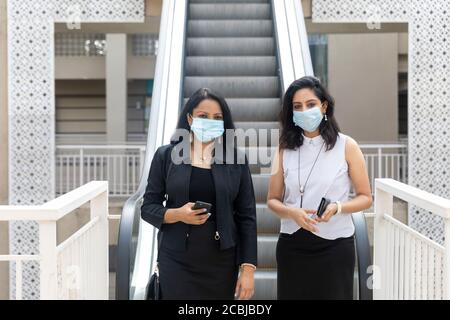 Deux femmes indiennes portent des masques de protection Covid-19 lorsqu'elles descendent un escalier mécanique dans un cadre urbain d'entreprise, Banque D'Images