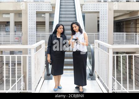 Deux femmes indiennes souriantes regardant dans un téléphone alors qu'elles descendaient un escalier mécanique dans un cadre urbain d'entreprise, Banque D'Images