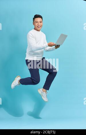 Image d'un jeune homme habillé debout sur fond bleu en utilisant un ordinateur portable tout en sautant. Banque D'Images