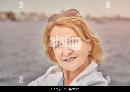 Portrait d'une femme blanche de race blanche d'âge attrayante et charmante, âgée d'environ 62 ans, avec des cheveux blonds et des lunettes de soleil. La dame retraité a un repos dans la cour Banque D'Images