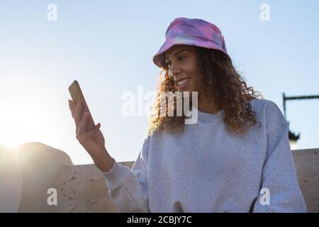 Portrait d'une jeune femme noire attrayante assise et parlant avec le téléphone mobile à l'extérieur avec son masque éteint. Communication de concept Banque D'Images