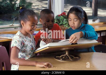 Austin, Texas, États-Unis. 1990: Trio ethniquement mixte d'élèves de l'école primaire recherchant des mots dans un grand dictionnaire non abrégé à la bibliothèque de l'école. Autorisation du modèle. ©Bob Daemmrich Banque D'Images