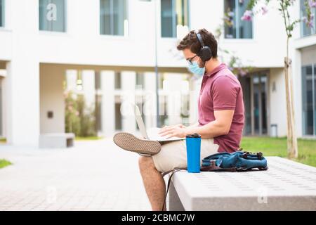 Le beau homme en masque de protection travaille avec un ordinateur portable sur le campus, pendant le concept de pandémie de coronavirus Banque D'Images