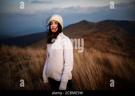 Jeune belle femme dans un chandail blanc se dresse au sommet d'une montagne et apprécie le paysage. Jeune femme debout sur le bord de la falaise et regardant un ciel Banque D'Images