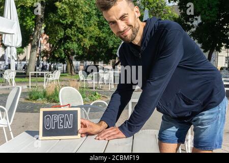 Garçon avec panneau ouvert sur la table café en plein air. Affiche de maintien du barista souriant ouverte. Le propriétaire du café accueille les clients. Rouvrir pour l'entreprise. Banque D'Images