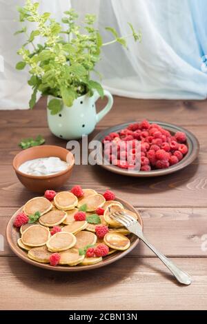 Crêpe de céréales sur la fourchette. Framboises fraîches mûres rouges dans une assiette brune et un bouquet de Melissa dans une tasse, yaourt sur une table en bois sombre près du vent Banque D'Images