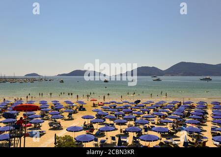 Vue panoramique sur une plage de sable avec des rangées de parasols, vacanciers sur la rive et le promontoire de Porto Venere en été, Lerici, Ligurie, Italie Banque D'Images