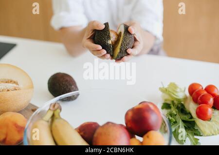 La moitié d'avocat parfaitement mûr chez la femme mains sur fond de fruits et légumes frais dans la cuisine moderne blanche. Concept d'alimentation saine. Cuisine maison Banque D'Images