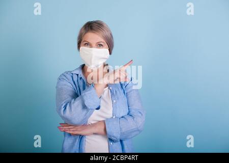 La femme âgée de race blanche avec un masque médical pointe vers le espace libre près de sa publicité quelque chose sur un studio bleu mur avec espace libre Banque D'Images