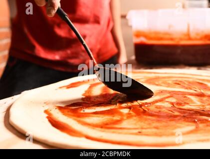 Sauce mettre sur la pizza italienne avec une cuillère. Processus de fabrication de pizza : le chef en tablier rouge met de la sauce. Pâte à pizza recouverte de sauce tomate rouge sur fond flou. Cuisine maison et concept de cuisine italienne. Banque D'Images