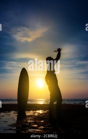 Silhouette de surf homme stand avec une planche de surf boire l'eau de la bouteille. Surf sur la plage au coucher du soleil. Sports nautiques en plein air aventure style de vie. Sports d'été Banque D'Images