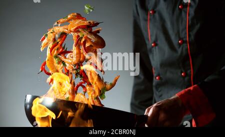 Gros plan du chef qui lance des crevettes dans un poêle à wok au feu. Préparation de plats asiatiques frais sur fond sombre. Banque D'Images