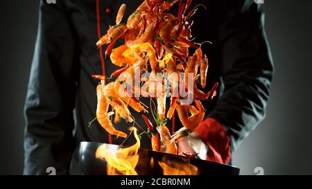 Gros plan du chef qui lance des crevettes dans un poêle à wok au feu. Préparation de plats asiatiques frais sur fond sombre. Banque D'Images