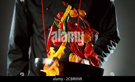 Gros plan du chef qui lance le mélange de piments du wok dans le feu. Préparation de plats asiatiques frais sur fond sombre. Banque D'Images