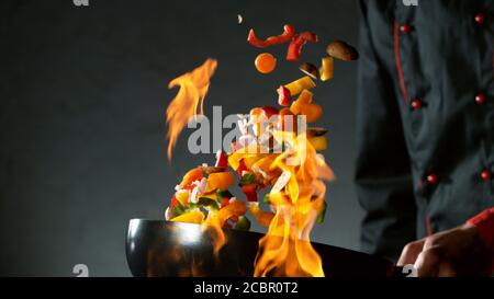 Gros plan du chef qui lance le mélange de légumes dans un poêle à wok au feu. Préparation de plats asiatiques frais sur fond sombre. Banque D'Images