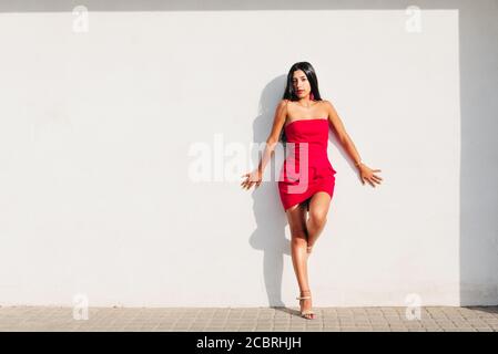 portrait de jeune fille latine élégante avec de longs cheveux de brunette et robe rouge sur le mur blanc avec une forte ombre, regardant l'appareil photo. espace de copie Banque D'Images