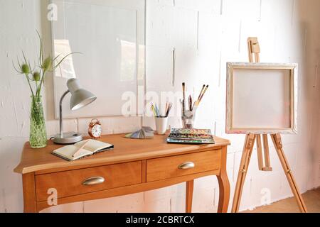 Petit lieu de travail d'un artiste avec des crayons, un carnet d'esquisses, une boîte d'aquarelles et des pinceaux sur une table et un chevalet contre un mur peint blanc rugueux, copie sp Banque D'Images