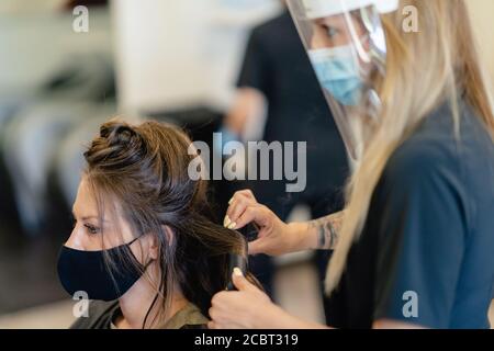 Coiffeur, protégé par un masque, peignant les cheveux de son client avec un fer à repasser dans un salon. Banque D'Images