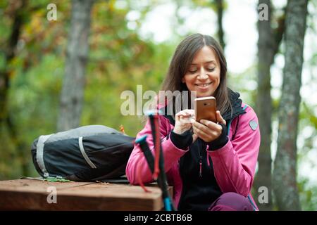 Une jeune fille de randonneurs se reposant sur un banc dans la forêt. Sac à dos avec veste rose tenant le téléphone cellulaire. Banque D'Images