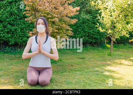 Jeune femme méditant à l'extérieur dans un parc ou un jardin avec masque chirurgical - concept de faire des exercices de yoga et de fitness pendant ou après le coronavirus Banque D'Images