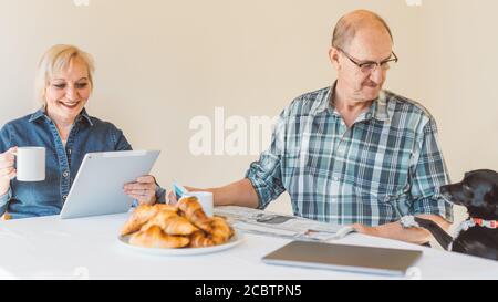 Un couple senior a lu des informations sur la tablette numérique et l'ancien style journal tout en prenant le petit déjeuner le matin - petit animal de compagnie (chien) demandant de la nourriture dur Banque D'Images