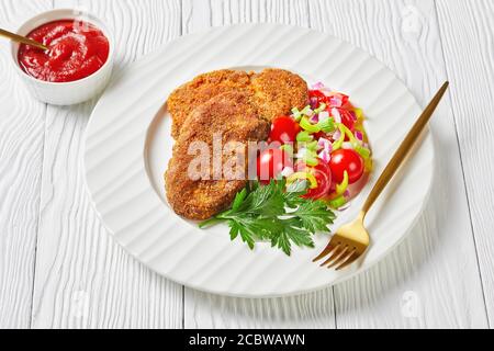 côtelettes d'agneau panées servies avec de la tomate, de l'oignon rouge, de la salade de poivron vert sur une assiette blanche sur une table en bois, vue panoramique Banque D'Images