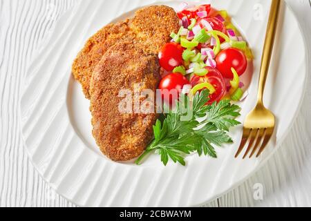 gros plan de côtelettes d'agneau panées servies avec de la tomate, de l'oignon rouge et de la salade de poivron vert sur une assiette blanche sur une table en bois Banque D'Images