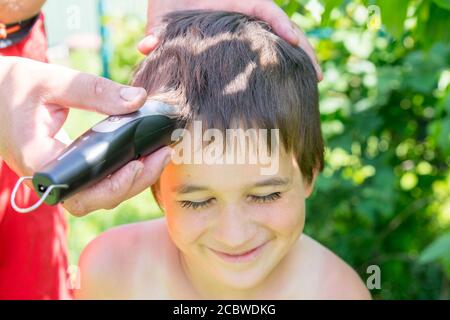 main tient une tondeuse à cheveux électrique et coupe les cheveux longs du garçon, barbershop à la maison, parent coupe les cheveux pendant que les coiffeurs sont fermés, rester à la maison Banque D'Images