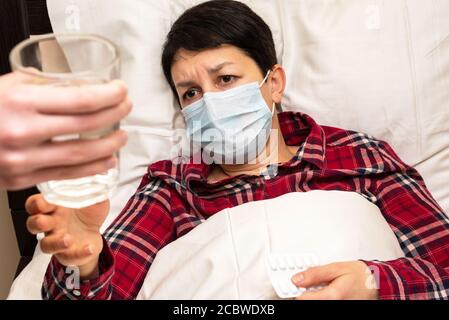 La femme malade dans un masque médical allongé sur le lit reçoit un verre d'eau pour prendre des pilules. Le concept de soins familiaux pour les malades Banque D'Images