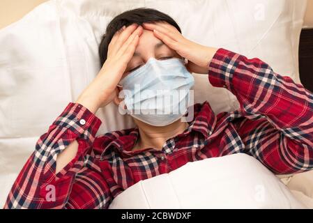 Femme malade dans un masque médical couché dans le lit souffrant de fièvre et de maux de tête. Brunette à poil court avec symptômes de covid-19 restant à la maison Banque D'Images