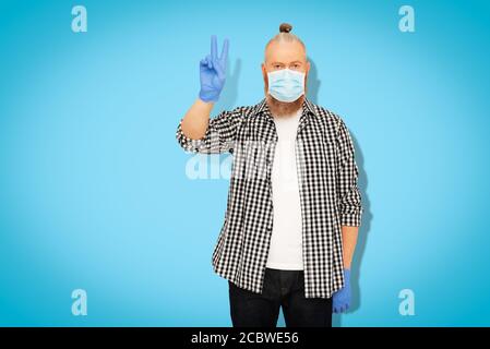 Homme barbu rouge dans un masque médical et des gants de protection montrant un geste de victoire sur fond bleu. Personne guérie de covid-19 Banque D'Images