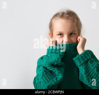 Adorable petite fille cachant la partie inférieure de son visage sous un col épais de pull chaud tricoté. Gros plan studio isolé sur espace de copie blanc Banque D'Images