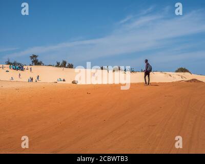 Sénégal, Afrique - 02 févr. 2019 : homme sur la route avec du sable rouge et blanc à la fin de la route Paris-Dakar passée et chidren d'école jouant sur le du Banque D'Images