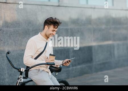 Un homme heureux en vêtements de travail tient une tasse, regarde la tablette et s'assoit sur un vélo dans la rue Banque D'Images