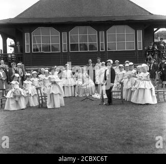 Années 1950, historique, la Reine Rose nouvellement couronnée assise dans sa robe avec d'autres filles dans leurs costumes et regardé par les gens locaux sur le terrain de jeu à Farnworth, Bolton, Lancashire, Angleterre, Royaume-Uni. Datant des années 1880, le festival annuel de la Reine des roses, qui a lieu en juin, est devenu un événement annuel majeur dans de nombreuses villes et villages à travers le Royaume-Uni, en particulier dans le Lancashire, connu sous le nom de comté de la rose rouge, à la suite des guerres des roses (1455-87). Banque D'Images