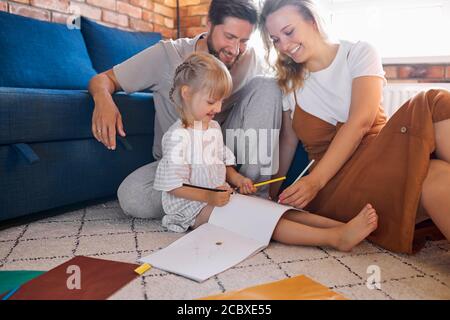 les parents heureux détendus s'assoient avec leur fille sur le sol, la petite fille caucasienne s'assoit à dessiner, avec des papiers à côté d'elle. à la maison Banque D'Images