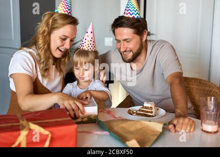 les parents heureux déballant les cadeaux d'anniversaire de leur belle fille, félicitent et célèbrent. à la maison dans la cuisine, dans les chapeaux de fête. concept de famille Banque D'Images