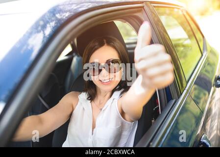 Une femme d'âge moyen et élégante portant des lunettes de soleil a mis sa main hors de la fenêtre de la voiture et a levé ses pouces. Banque D'Images