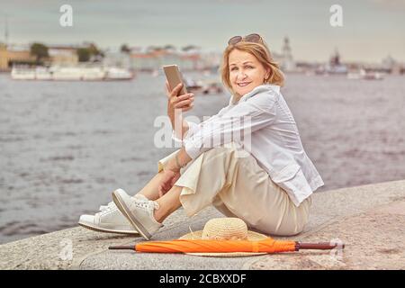 Belle femme d'âge caucasien active mature d'environ 62 ans avec les cheveux blonds et dans le tissu décontracté et élégant est assis Près de la rivière à Saint-Pétersbourg wi Banque D'Images