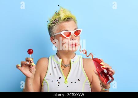 vieille femme élégante tenant le lollipop et une bouteille de jus et appréciant de le boire. gros plan portrait, plaisir, style de vie Banque D'Images