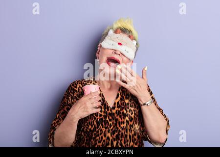 femme dormanteuse élégante senior dans le masque de sommeil bâilling, tenant une tasse de thé en plastique isolée sur bleu. gros plan portrait, studio tourné Banque D'Images