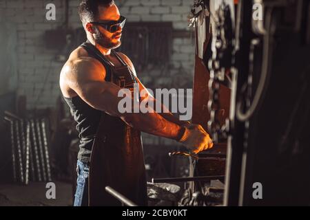 adulte musclé avec barbe portant un tablier en cuir travaillant dans l'atelier, chauffant. métal trempé dans le four Banque D'Images