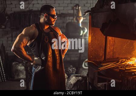 adulte musclé avec barbe portant un tablier en cuir travaillant dans l'atelier, chauffant. métal trempé dans le four Banque D'Images