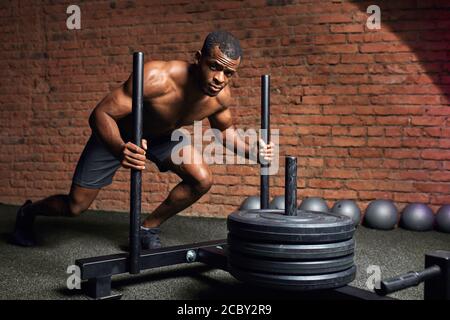 Bodybuilder mâle à la peau sombre exécutant un exercice de poussée de poids lourd de traîneau dans la salle de sport de cross-fit. Banque D'Images