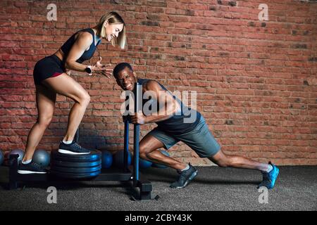 Un athlète africain qui travaille avec une machine de poussée de traîneau de poids lourd et une femme blonde européenne le soutient dans un studio de cross fit intérieur. Banque D'Images