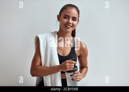 Positive souriante belle femme dans les vêtements de sport tenant une bouteille d'eau avec une serviette sur les épaules. Isolé dans le studio gris, salle Banque D'Images