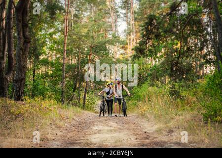 Les jeunes couples caucasiens de cyclistes marchent et poussent leurs VTT le long de la route forestière dans le parc. Week-end des sports actifs. Allure sportive et élégante Banque D'Images